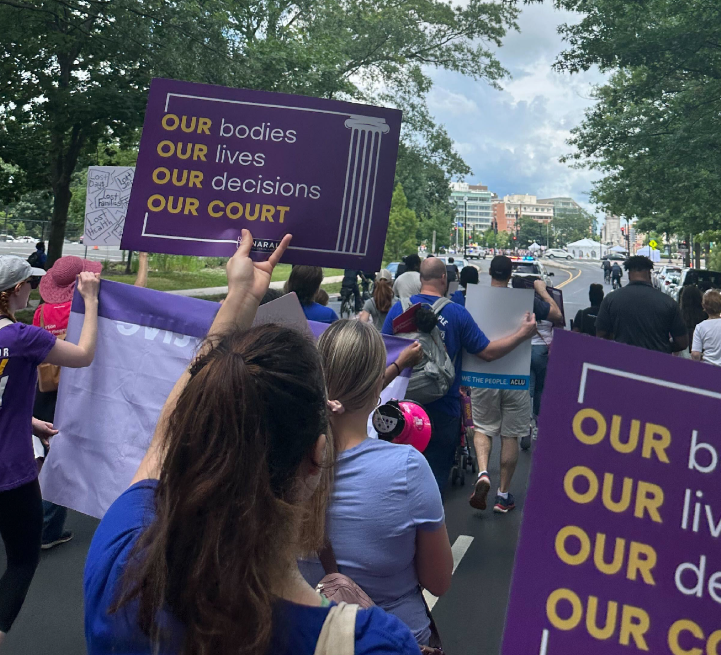 Image of a large group of protesters on the Dobbs anniversary, marching down a street in Washington, DC, with banners and signs, including one that says, “Our bodies, Our lives, Our decisions, Our Court.