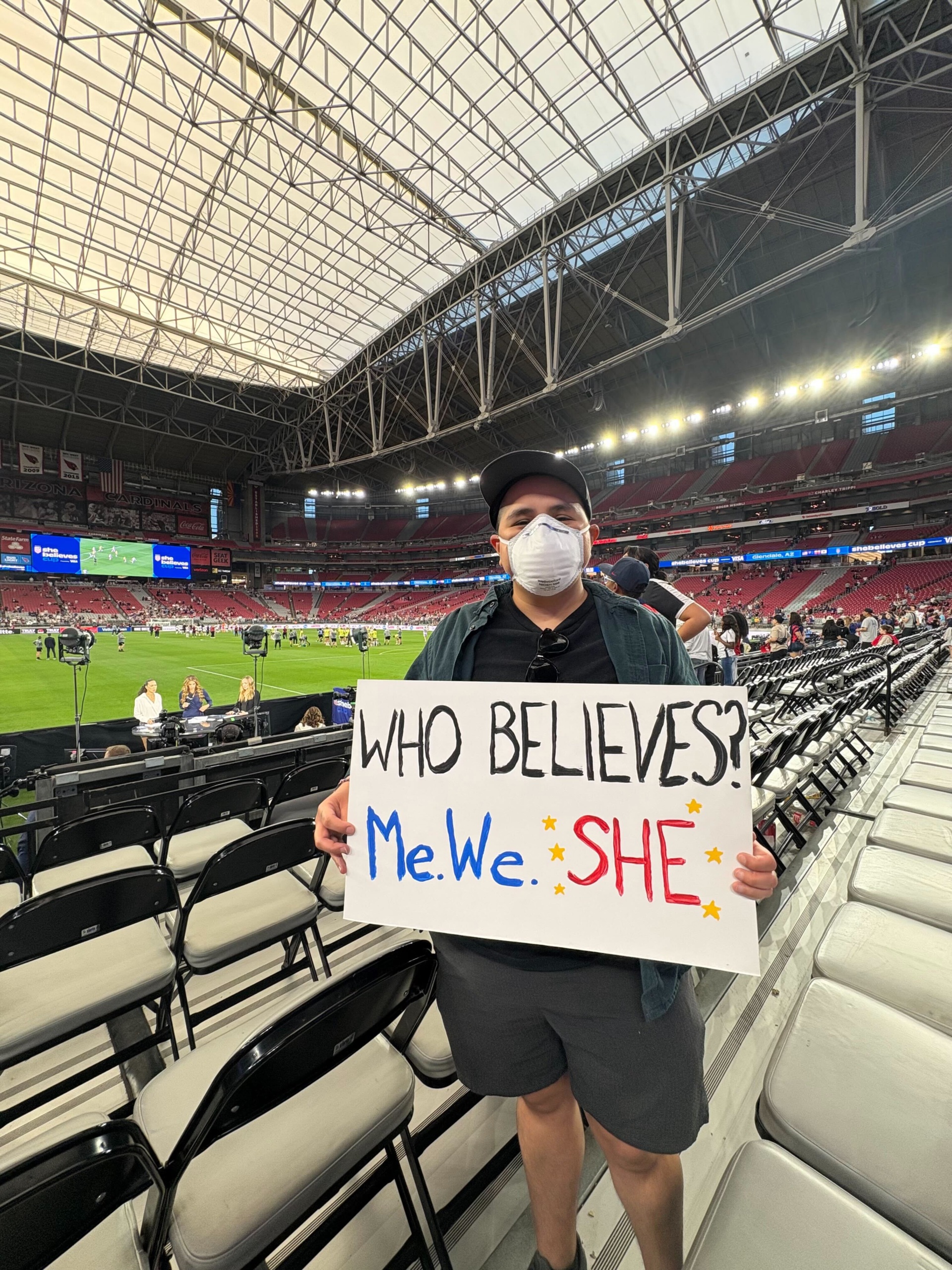 Kat M. at a soccer game holding a 'Who Believes? Me. We. She.' sign